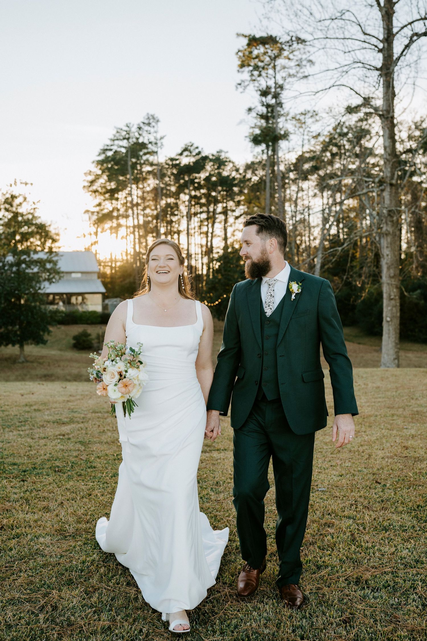 A couple shares a romantic moment in a field during golden hour with pine trees and a barn in the distance.