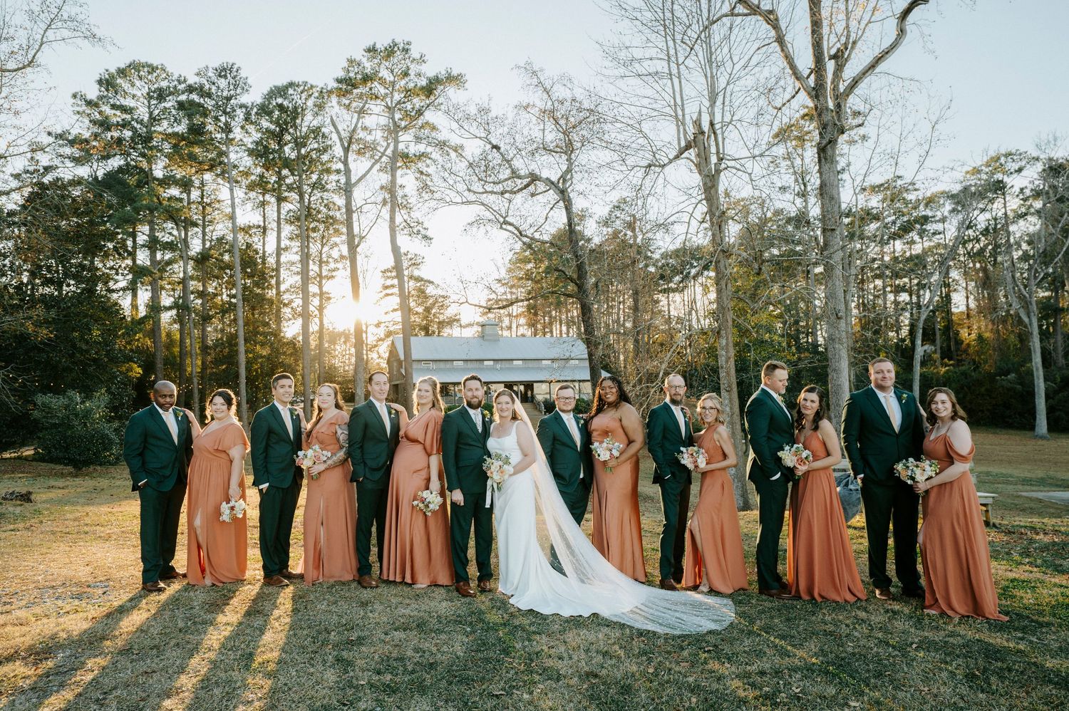 Wedding party in dusty rose bridesmaid dresses poses together in a sunlit forest setting during golden hour.