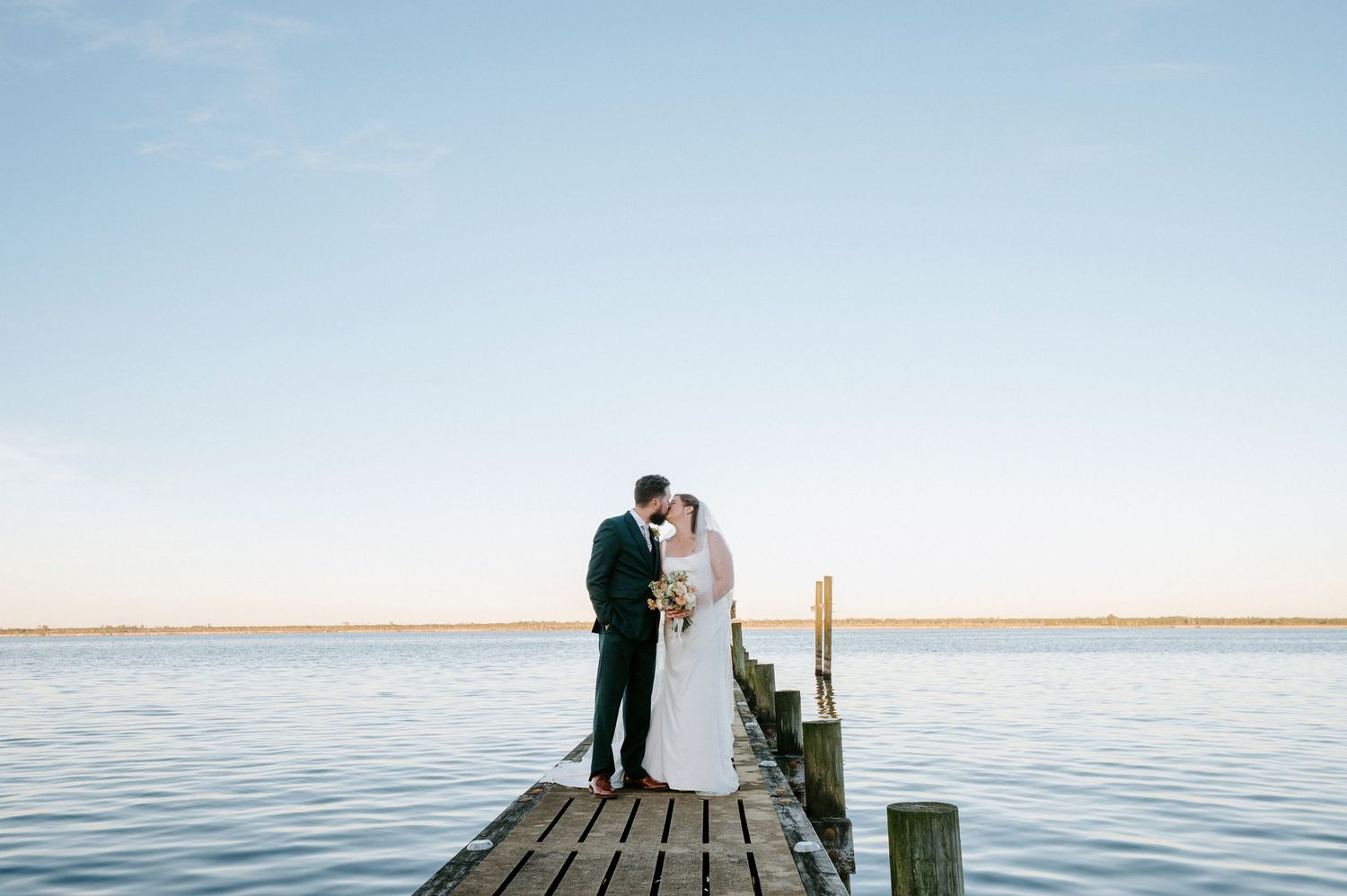 A couple stands on a wooden dock overlooking a serene lake during sunset.