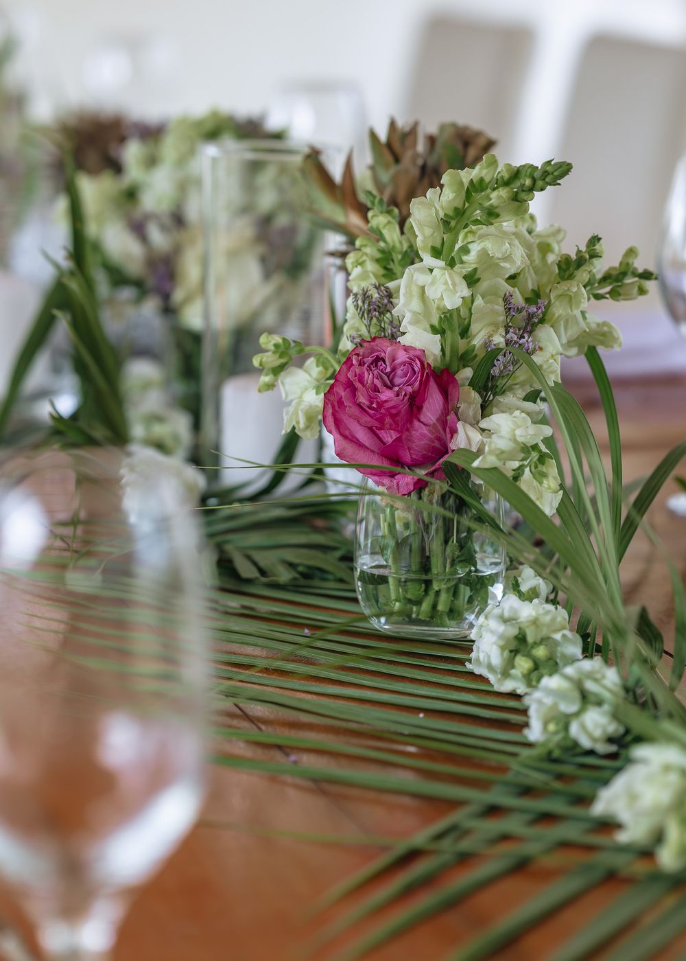 Pink rose and white floral centerpiece with greenery arranged on wooden table with wine glasses for elegant dining.