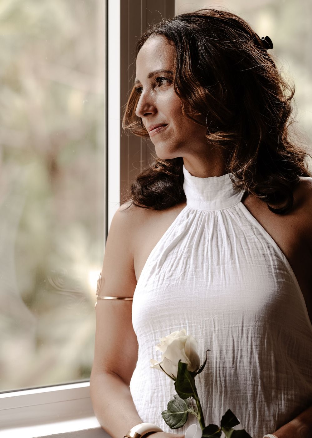 Woman in white sleeveless dress poses near window with soft natural lighting.