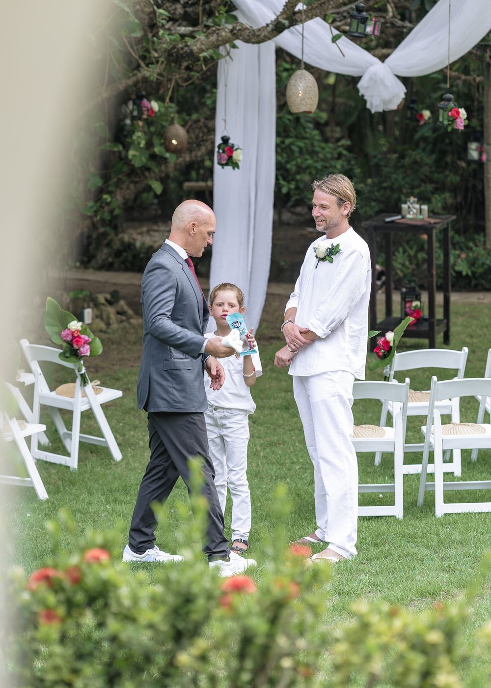 A wedding ceremony setup outdoors with white chairs and decorative draping between trees in a garden setting.