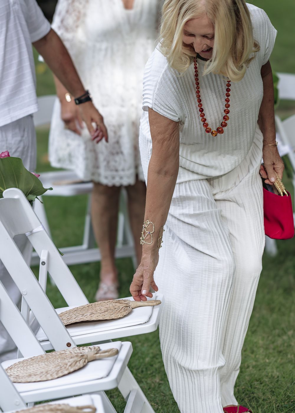 Woman in white dress placing items on white folding chairs on grass during outdoor event setup.