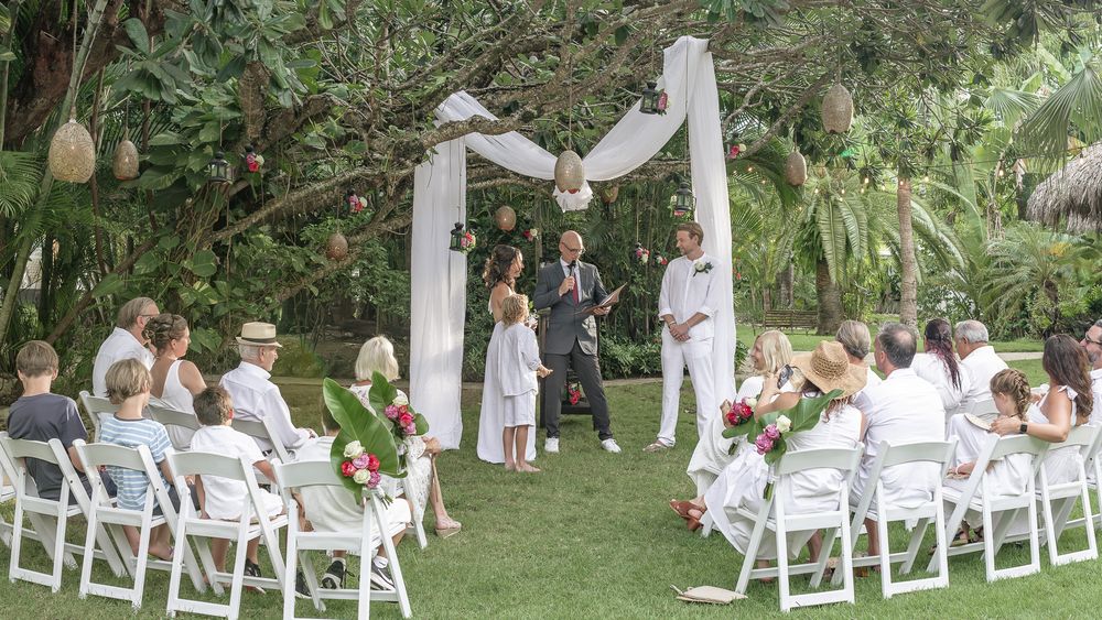 Panoramic view of an outdoor garden wedding ceremony with white chairs arranged in rows under lush green trees.