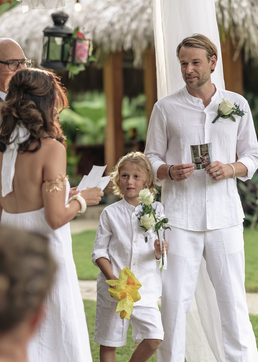 A wedding ceremony takes place outdoors under a white canopy with people in white attire exchanging vows.