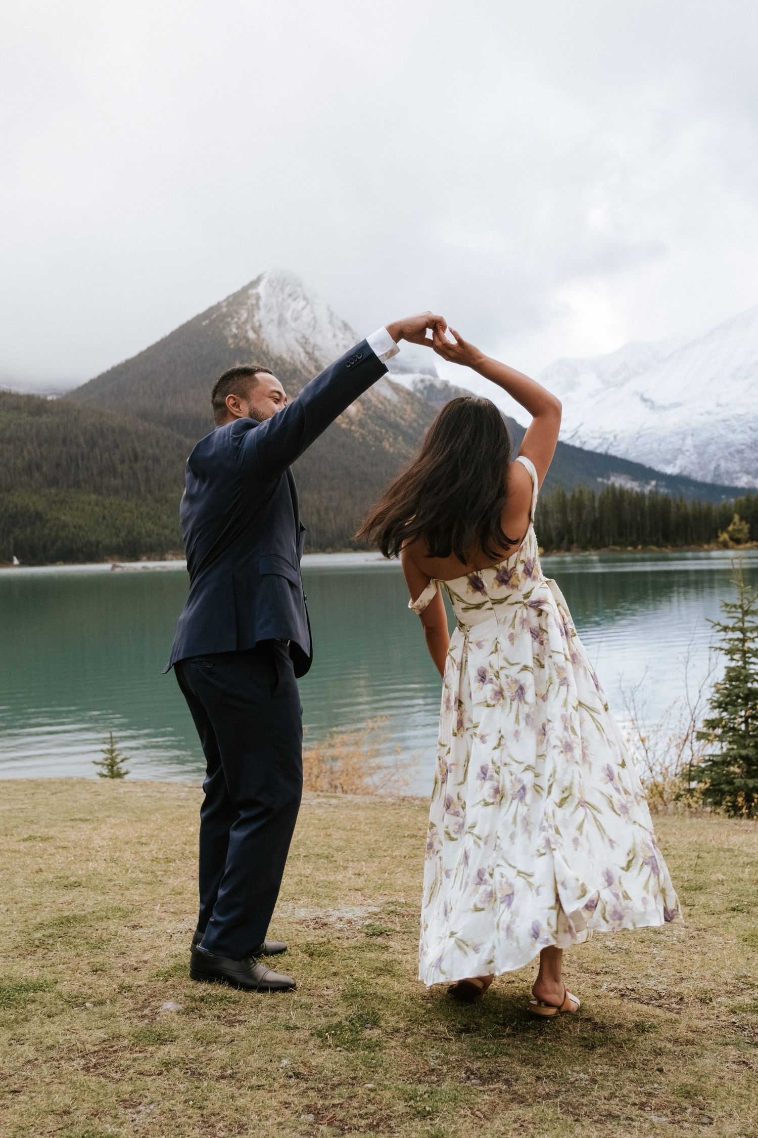 A couple in wedding attire dances playfully by a scenic mountain lake surrounded by evergreen trees and snow-capped peaks.