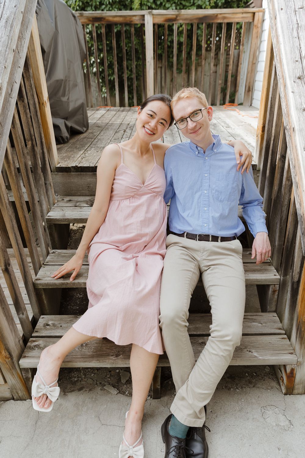 A couple sits together on wooden steps, with one wearing a pink dress and the other in khaki pants and blue shirt.