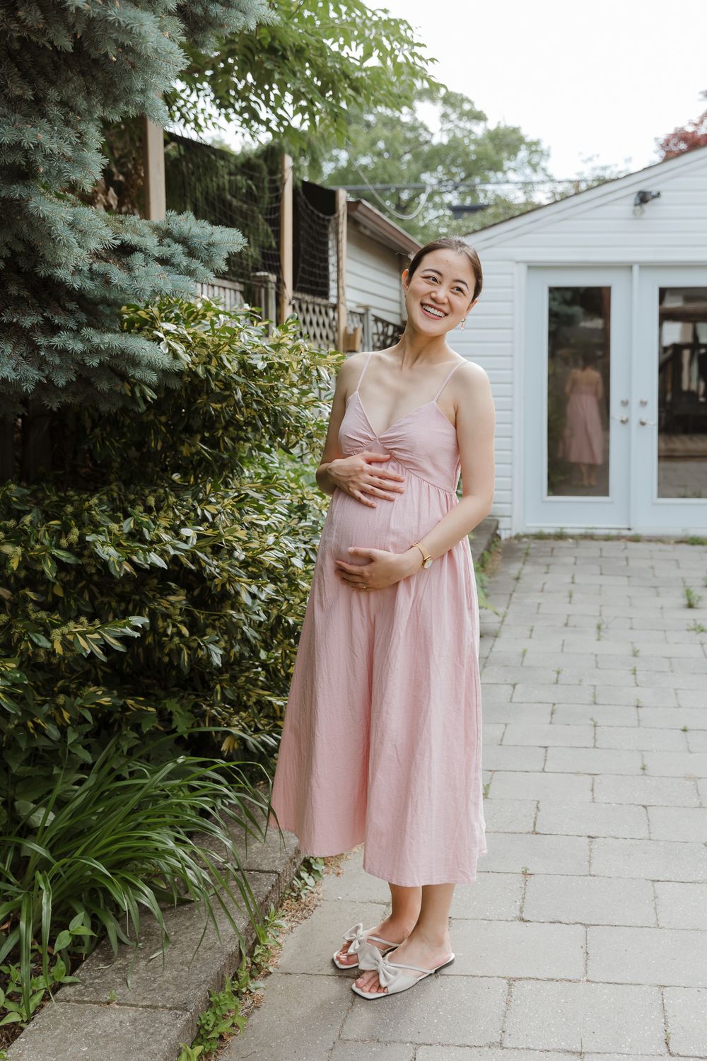 A person in a pink summer dress poses on stone pavers near a white house and garden.