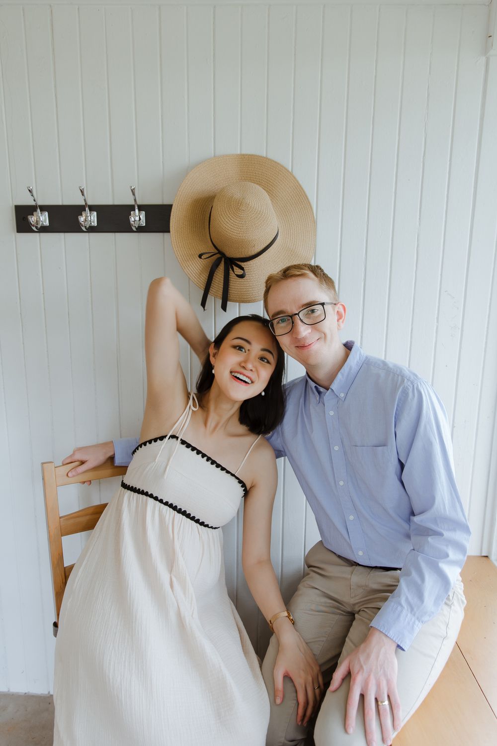 A couple poses together indoors against a white beadboard wall, with a straw hat hanging above on coat hooks.