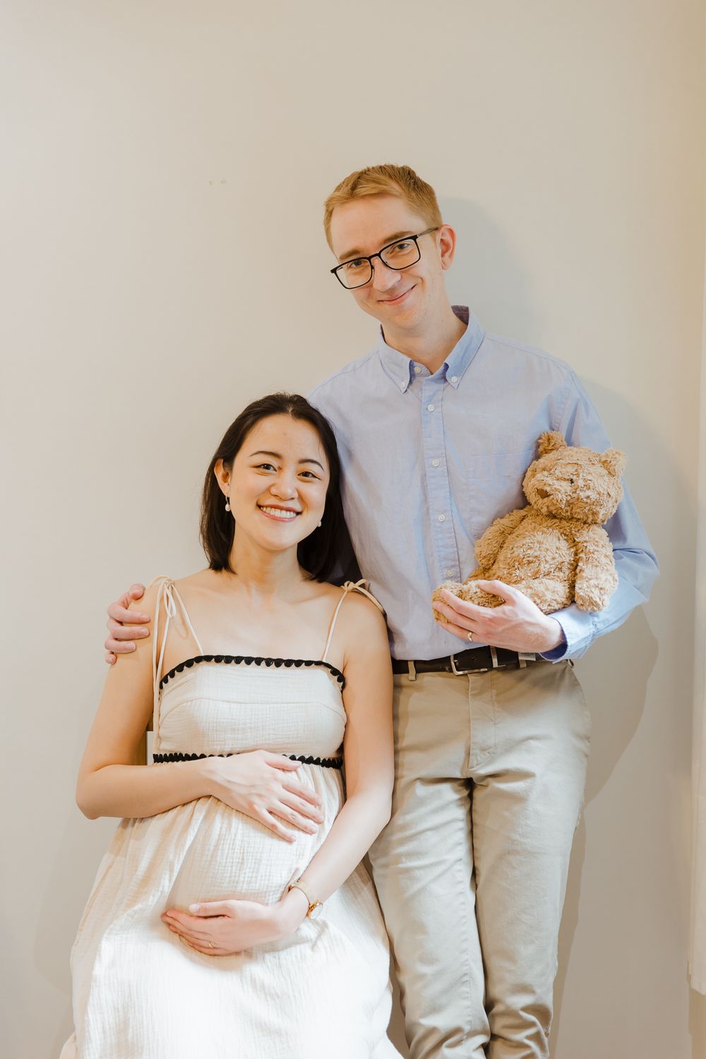 Expecting couple poses together against white wall holding teddy bear during maternity photo shoot.