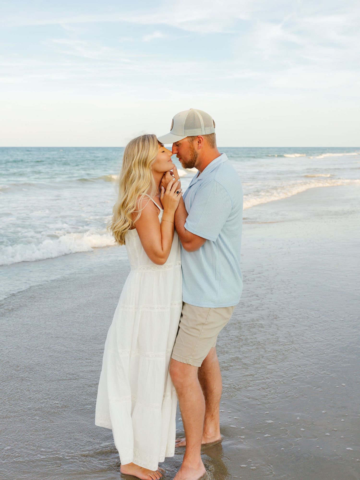 Romantic beach photo session with couple in white and beige outfits by ocean waves.