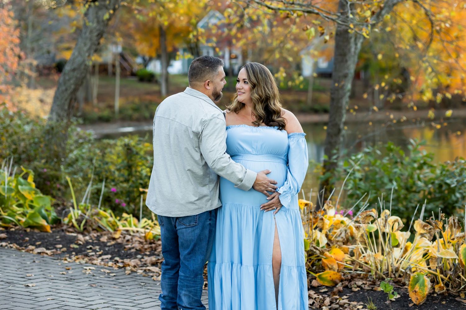 A couple in autumn scenery stands by a pond, with the person in a flowing light blue dress amid fall foliage.