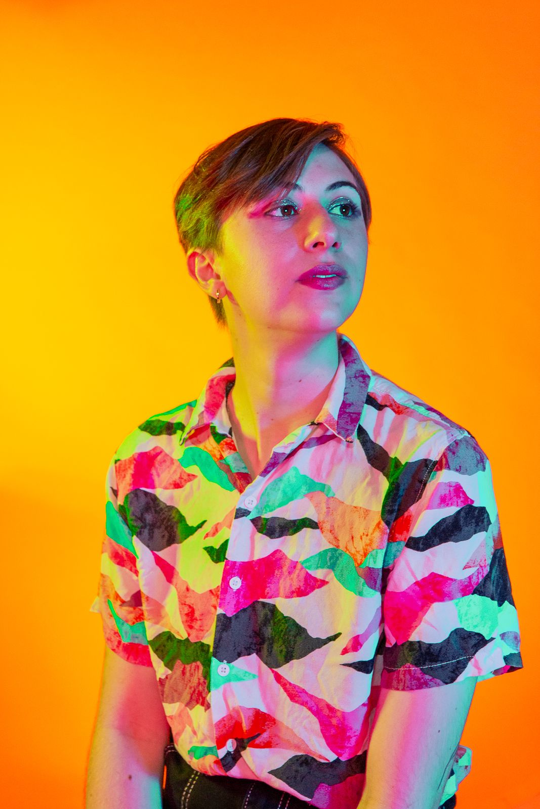 Portrait with colorful lighting wearing vibrant patterned shirt against orange backdrop.