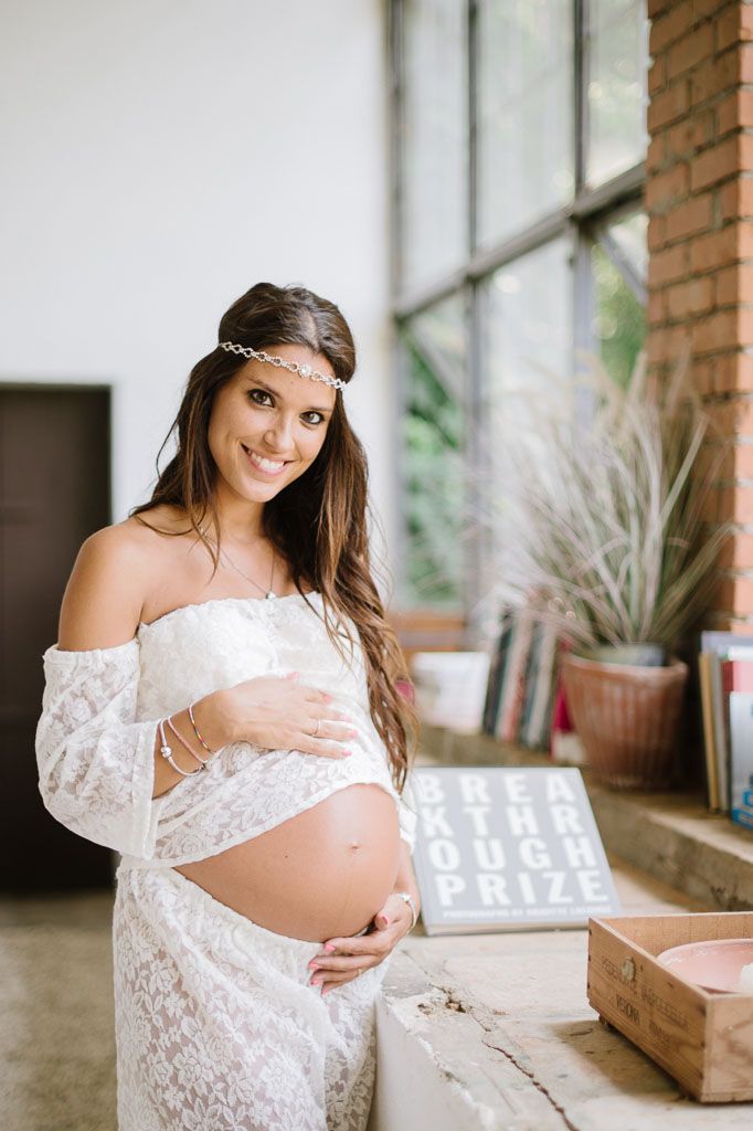 Bohemian maternity portrait in white lace dress near exposed brick and window.