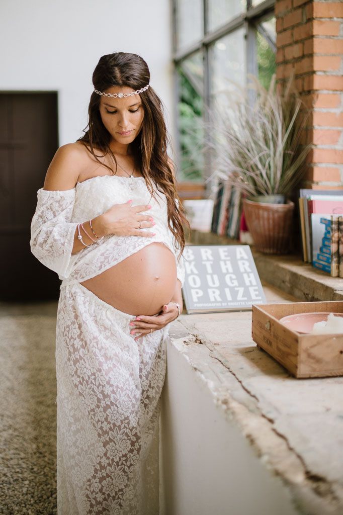 Artistic maternity photo with white lace dress against industrial brick wall setting.
