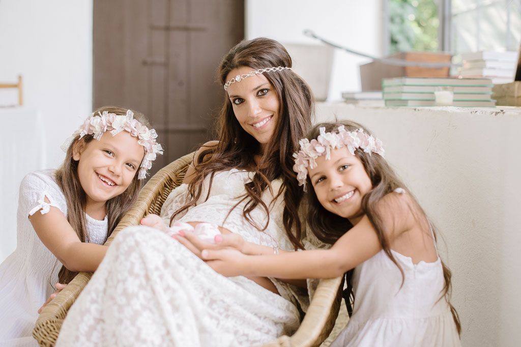Three figures in white dresses and floral headpieces share a tender moment together on a couch.