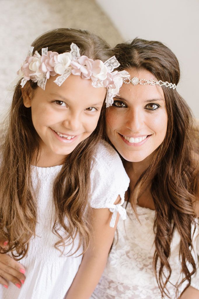 Two figures wearing white dresses and delicate floral crowns pose together for a portrait.