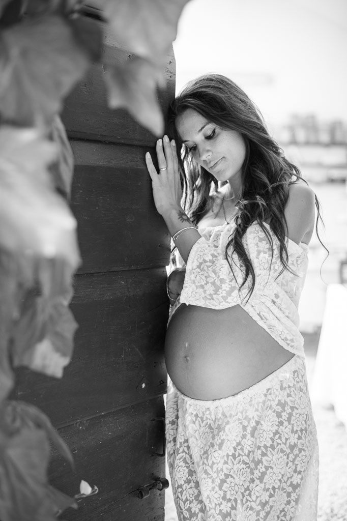 Woman in white lace dress rests against wall during maternity photoshoot in black and white.