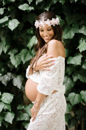 Expectant mother in white lace dress and flower crown smiles during outdoor photoshoot.