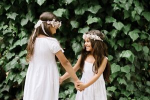 Two young figures in white dresses and floral crowns hold hands against ivy wall.