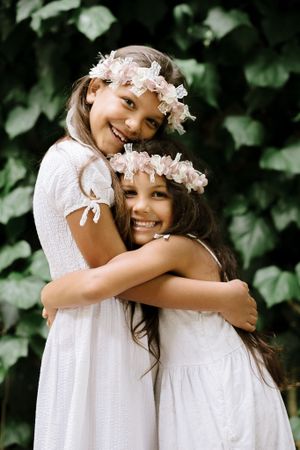 Two figures in white dresses and pink floral crowns share joyful embrace.