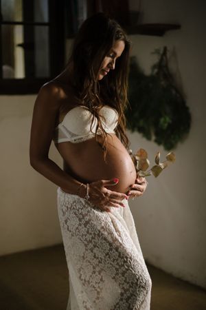 Woman in white lace dress cradles baby bump in moody indoor maternity portrait.
