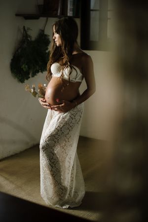 Pregnant woman in white lace dress poses in soft natural light for intimate maternity portrait.