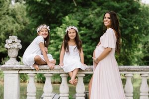 Outdoor maternity photo with children sitting on stone balustrade in garden setting.