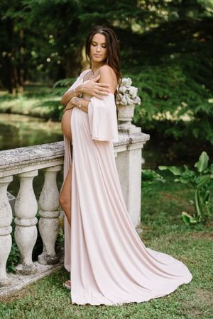 Ethereal maternity portrait of woman in flowing pink dress by stone balustrade.