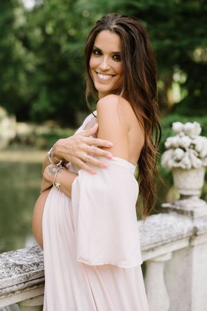 Joyful maternity portrait of woman in pink dress leaning against stone railing outdoors.