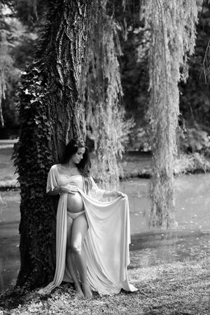 Artistic black and white maternity photo featuring a flowing white dress against large tree trunks in a natural setting.