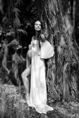 Ethereal black and white maternity portrait against textured tree bark in a woodland setting.