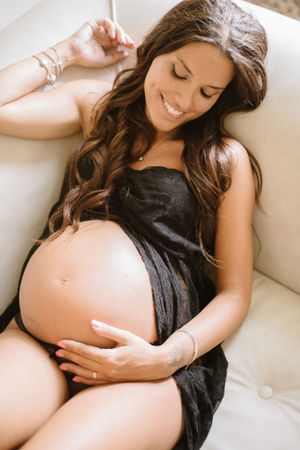 Expectant mother in black dress reclining on white couch with natural lighting.
