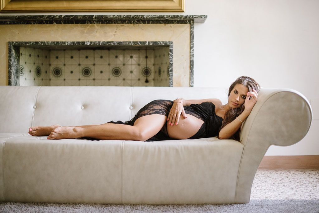 A dramatic boudoir photo of someone reclining on a beige chaise lounge beneath an ornate mirror and frame.