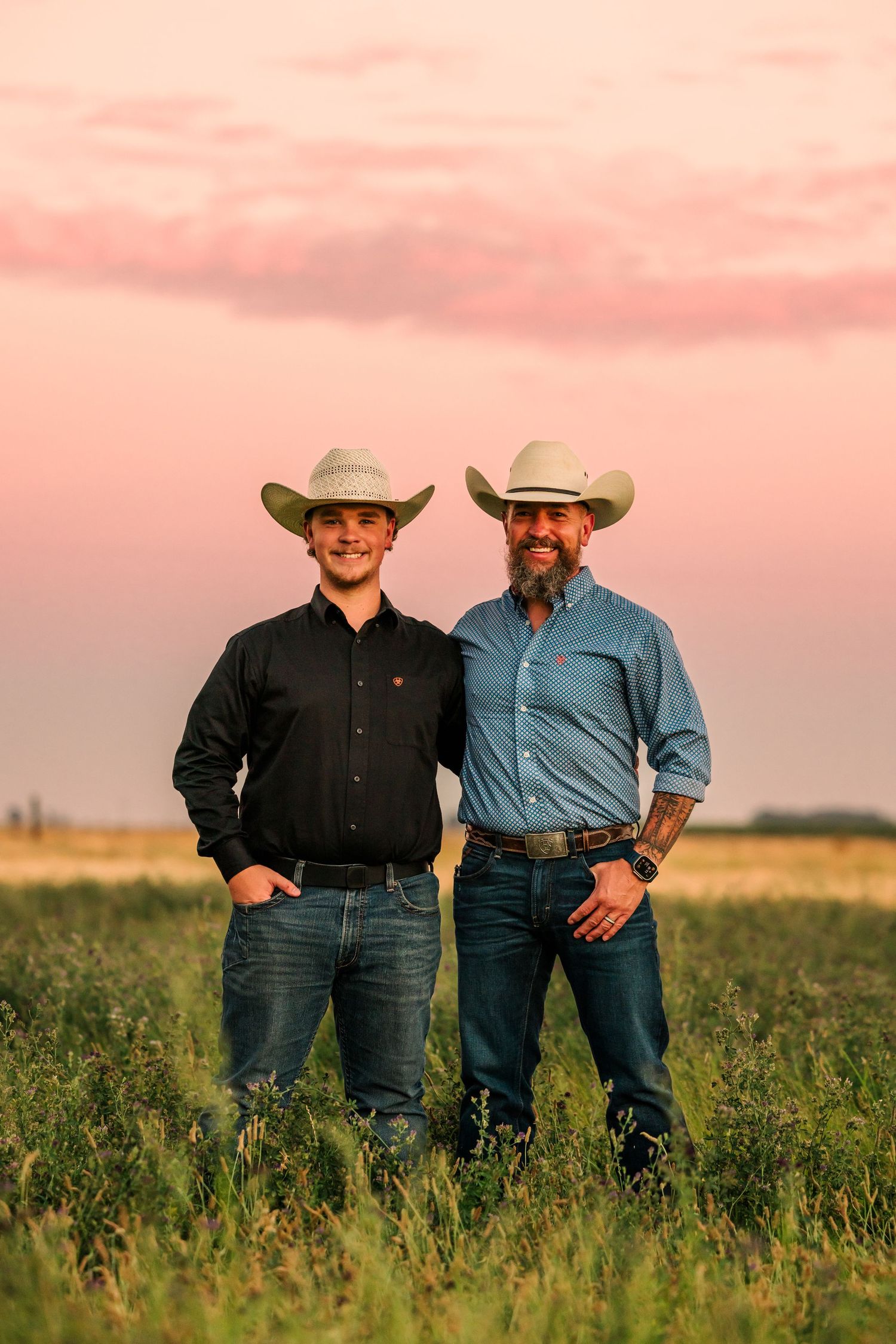 Two people in cowboy hats stand together in grassy field at sunset.