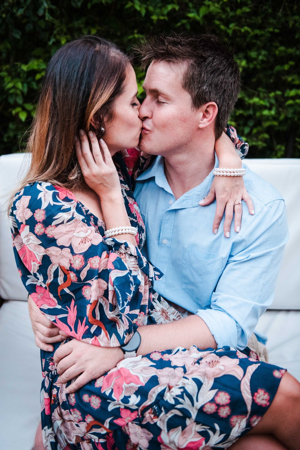 An image of a couple sitting together in a shaded forest area, looking at each other and kissing during their engagement shoot.