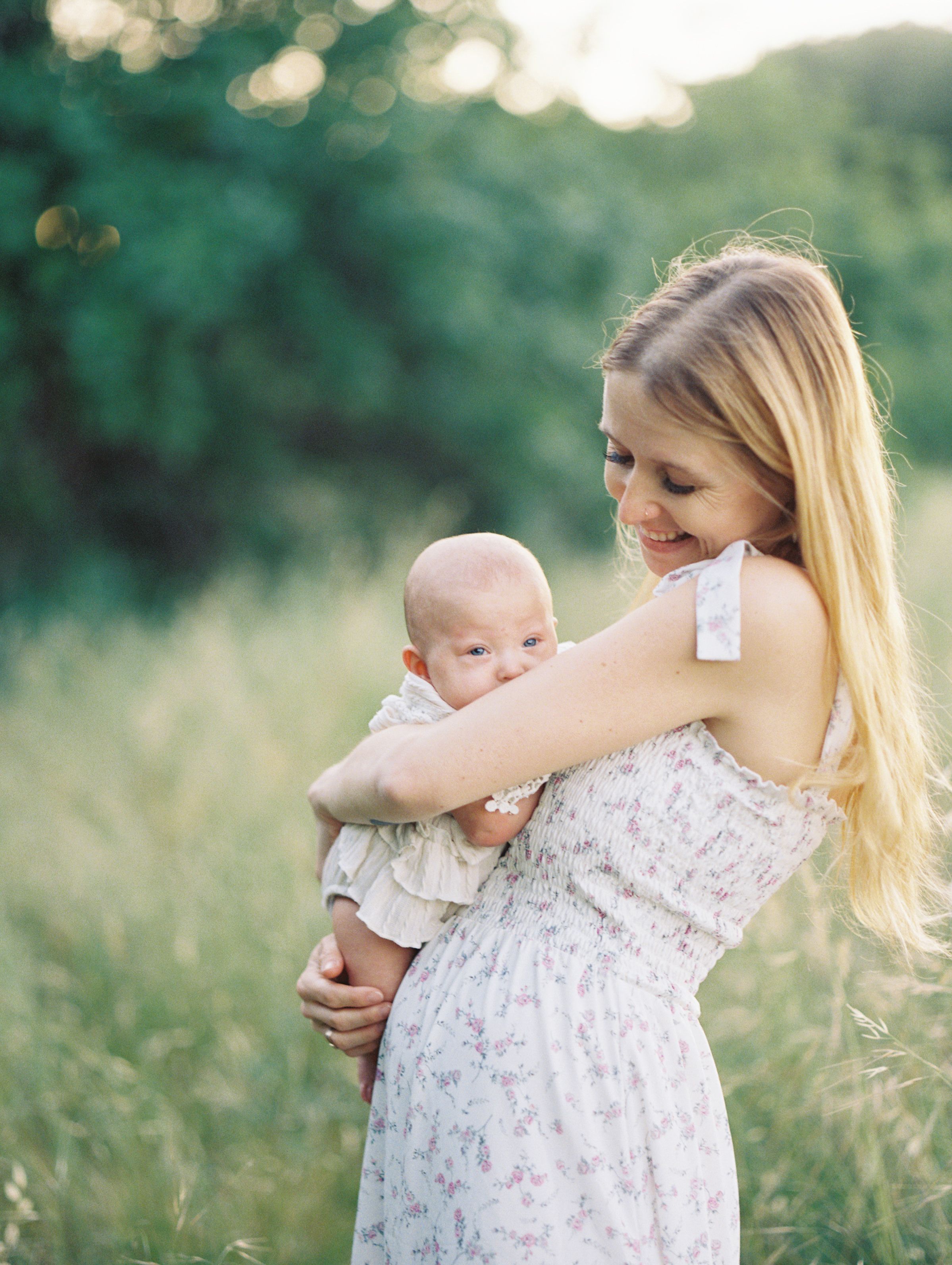 A smiling blonde mother in a floral dress tenderly holds her newborn baby in a sunlit green meadow.