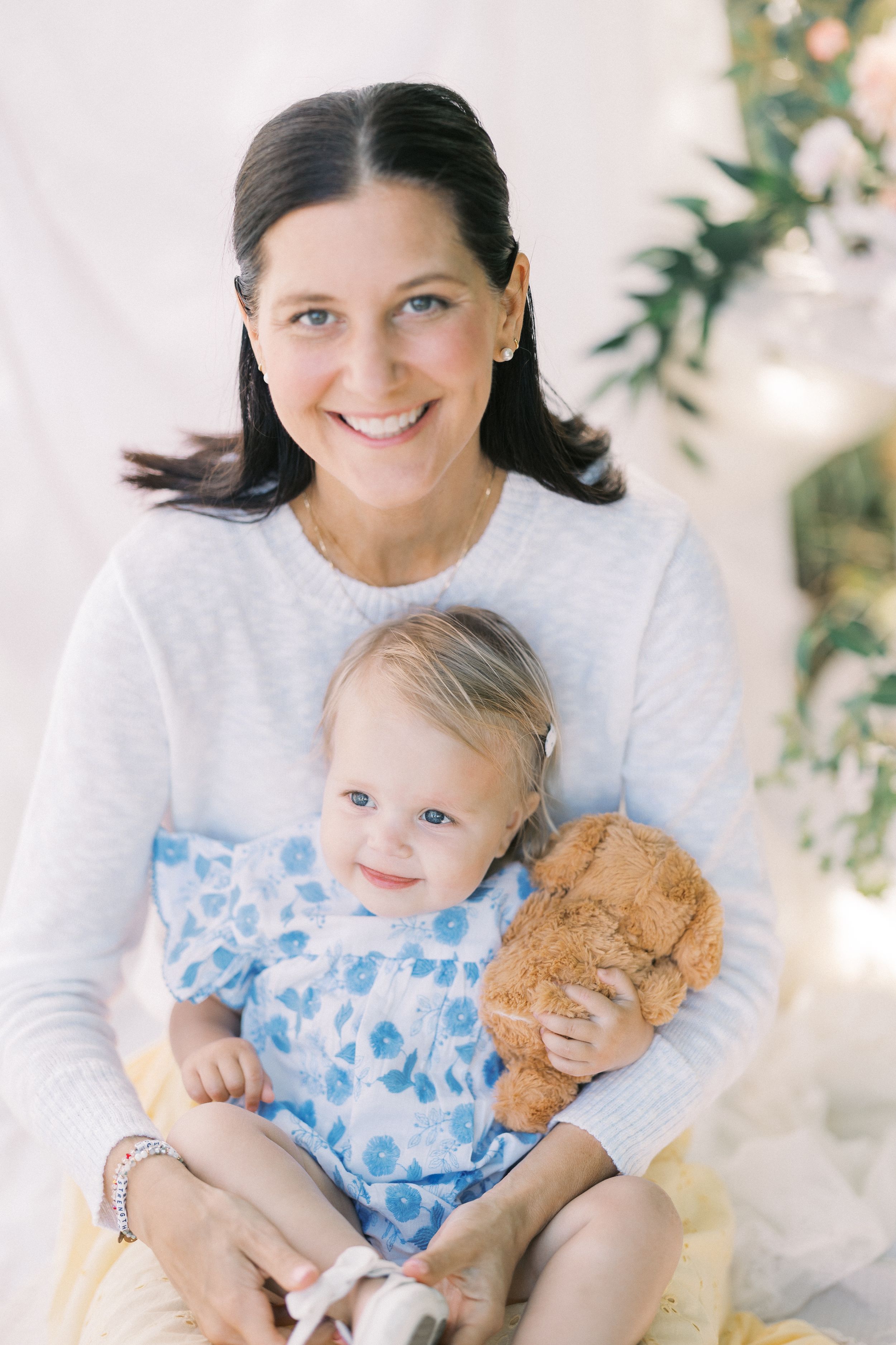 A smiling dark-haired woman in a white sweater holds a toddler girl in a blue floral dress clutching a teddy bear.