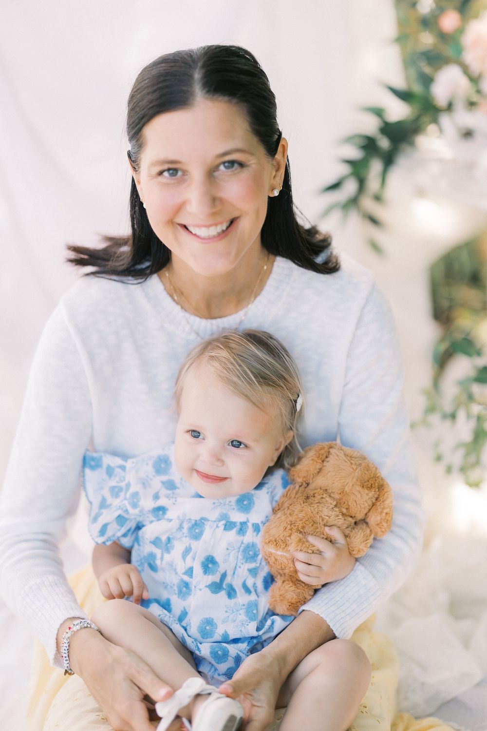 A smiling dark-haired woman in a white sweater holds a toddler girl in a blue floral dress clutching a teddy bear.
