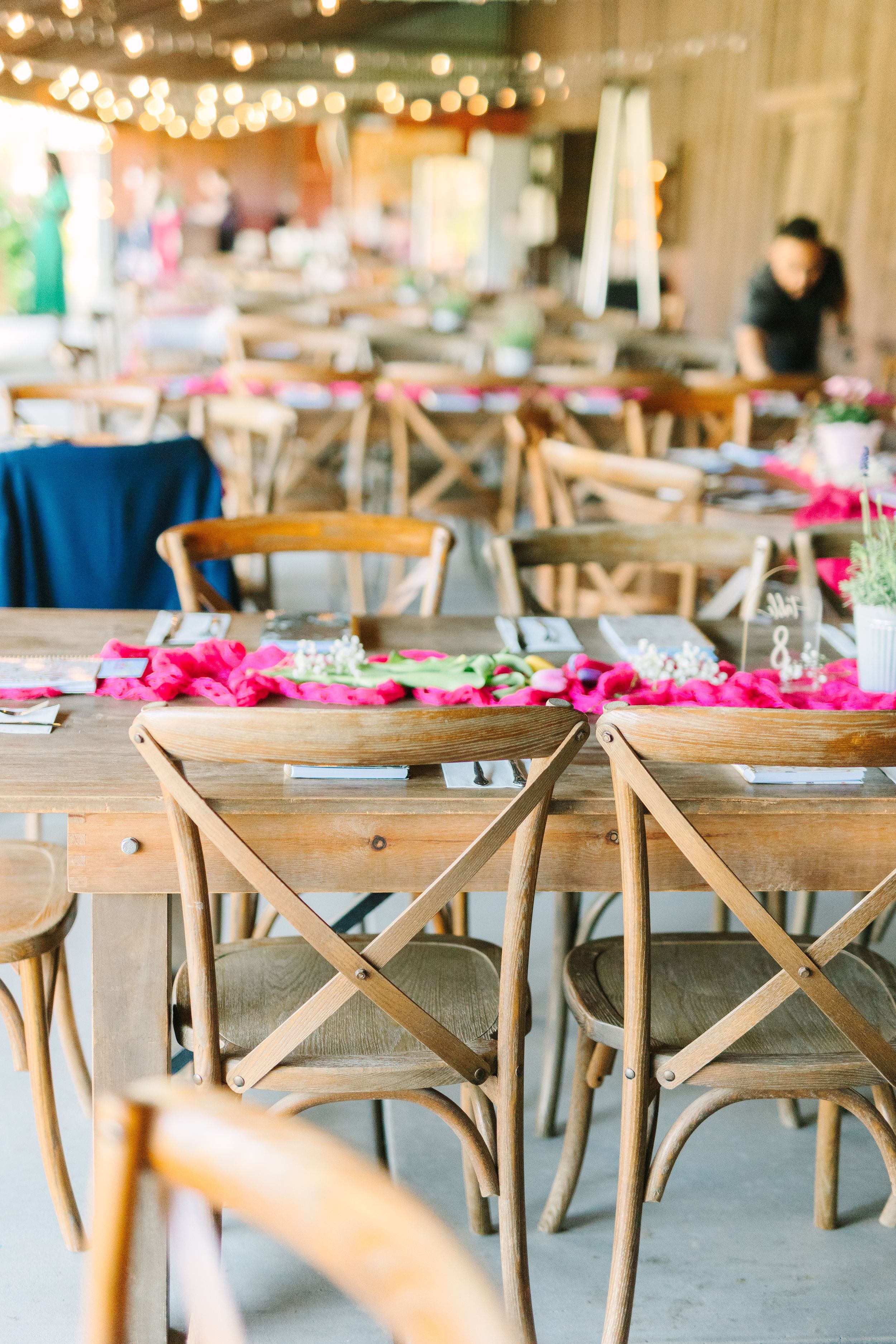 Rustic wooden farm tables with pink floral runners and cross-back chairs set for an elegant outdoor wedding reception.