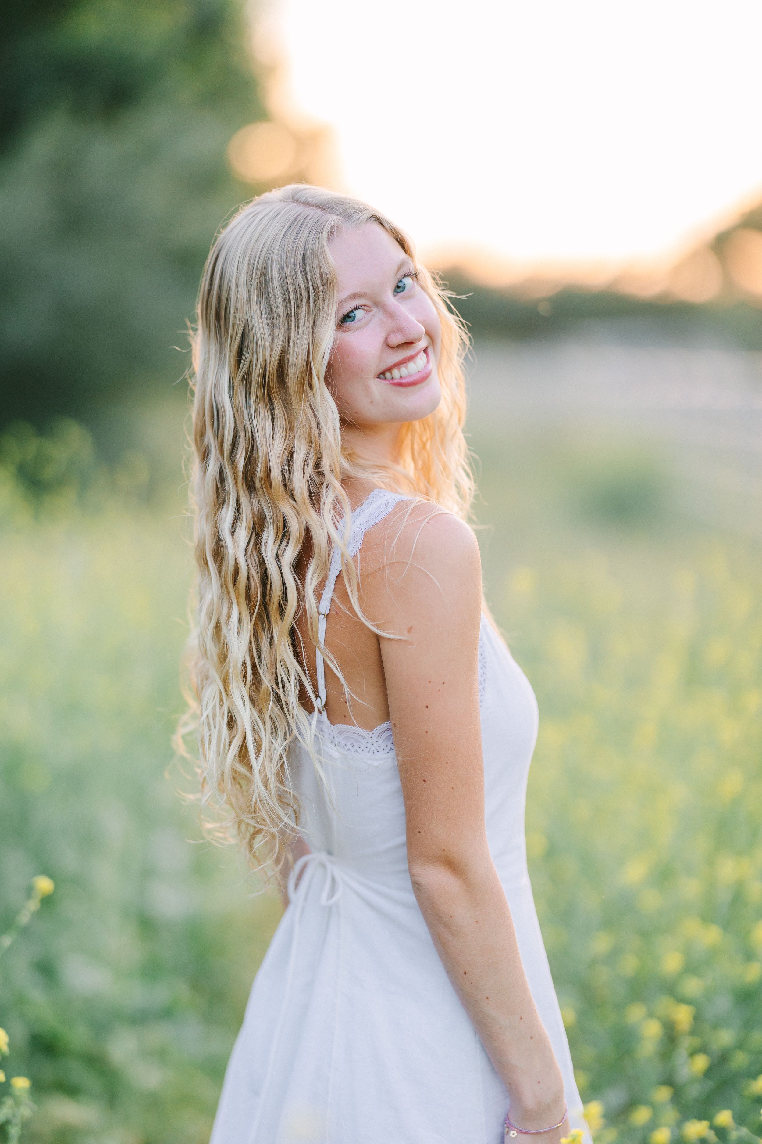 Smiling blonde woman in white dress standing in sunny yellow wildflower field with long wavy hair.