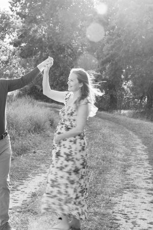 Black and white photo of a pregnant woman dancing with a man on a rural path, sunlight filtering through trees.
