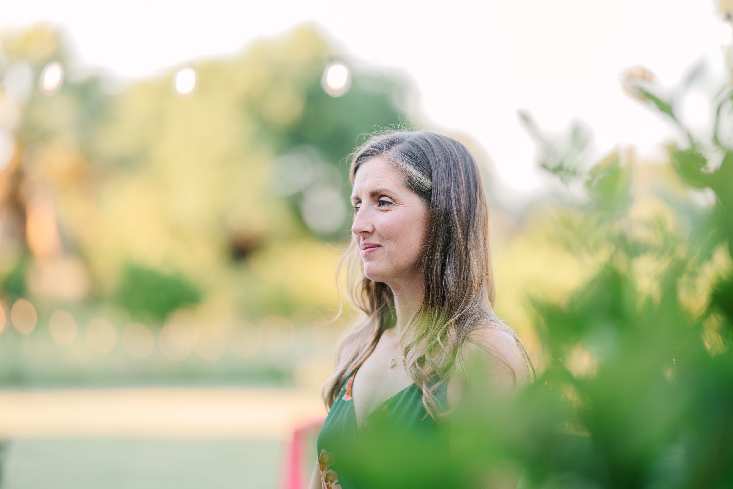 A young woman with long hair in a green dress smiles outdoors, surrounded by lush greenery in golden light.