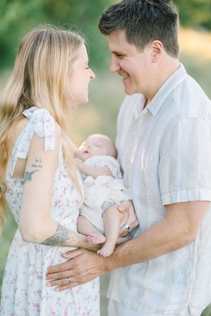A smiling couple in soft pastel outfits lovingly gazes at their newborn baby outdoors in natural light.