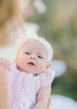 Newborn baby girl in pink dress and white lace headband being held up, looking alert with bright eyes.