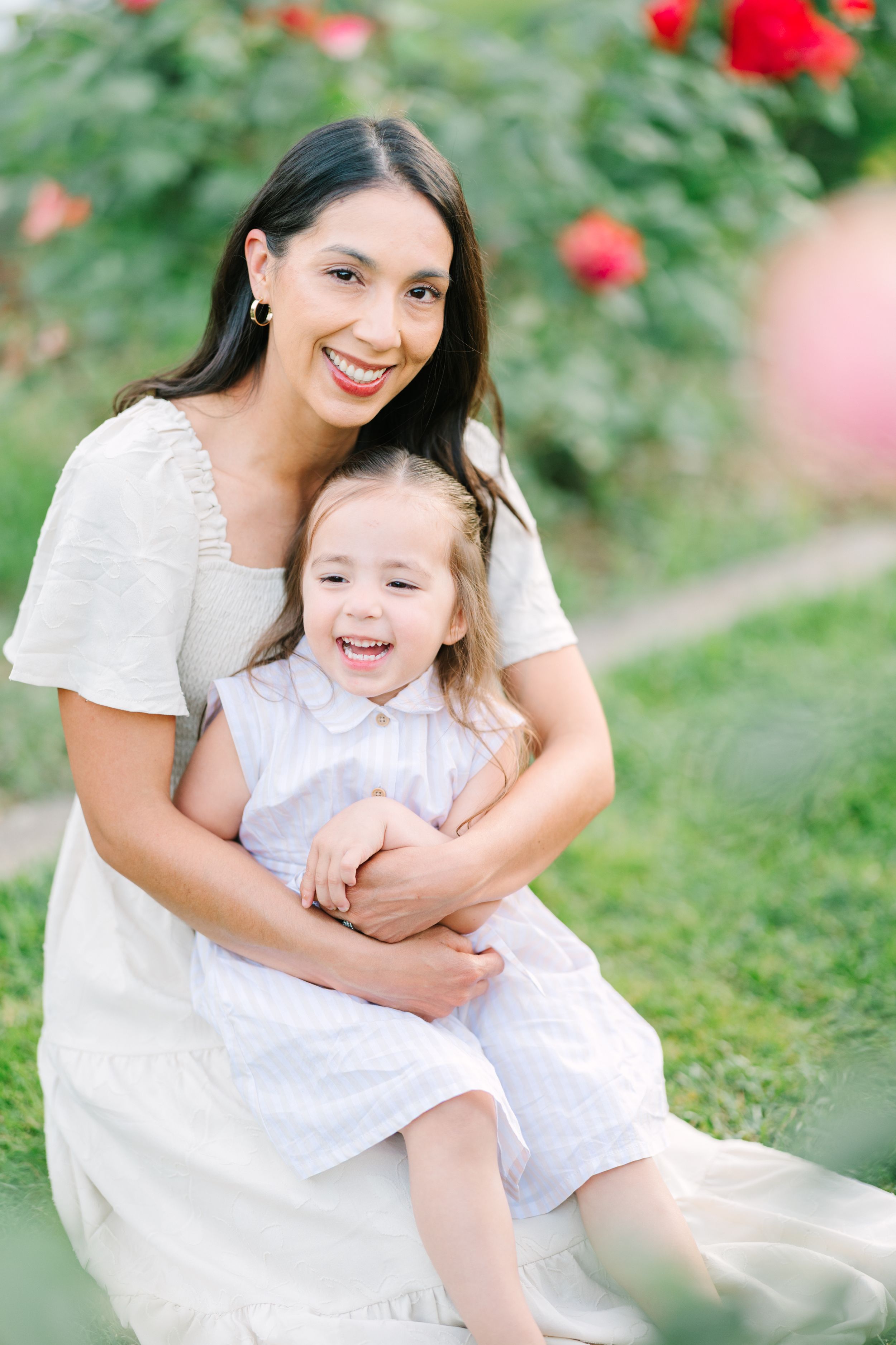 Smiling mother in white dress hugging laughing toddler girl outdoors near red roses.