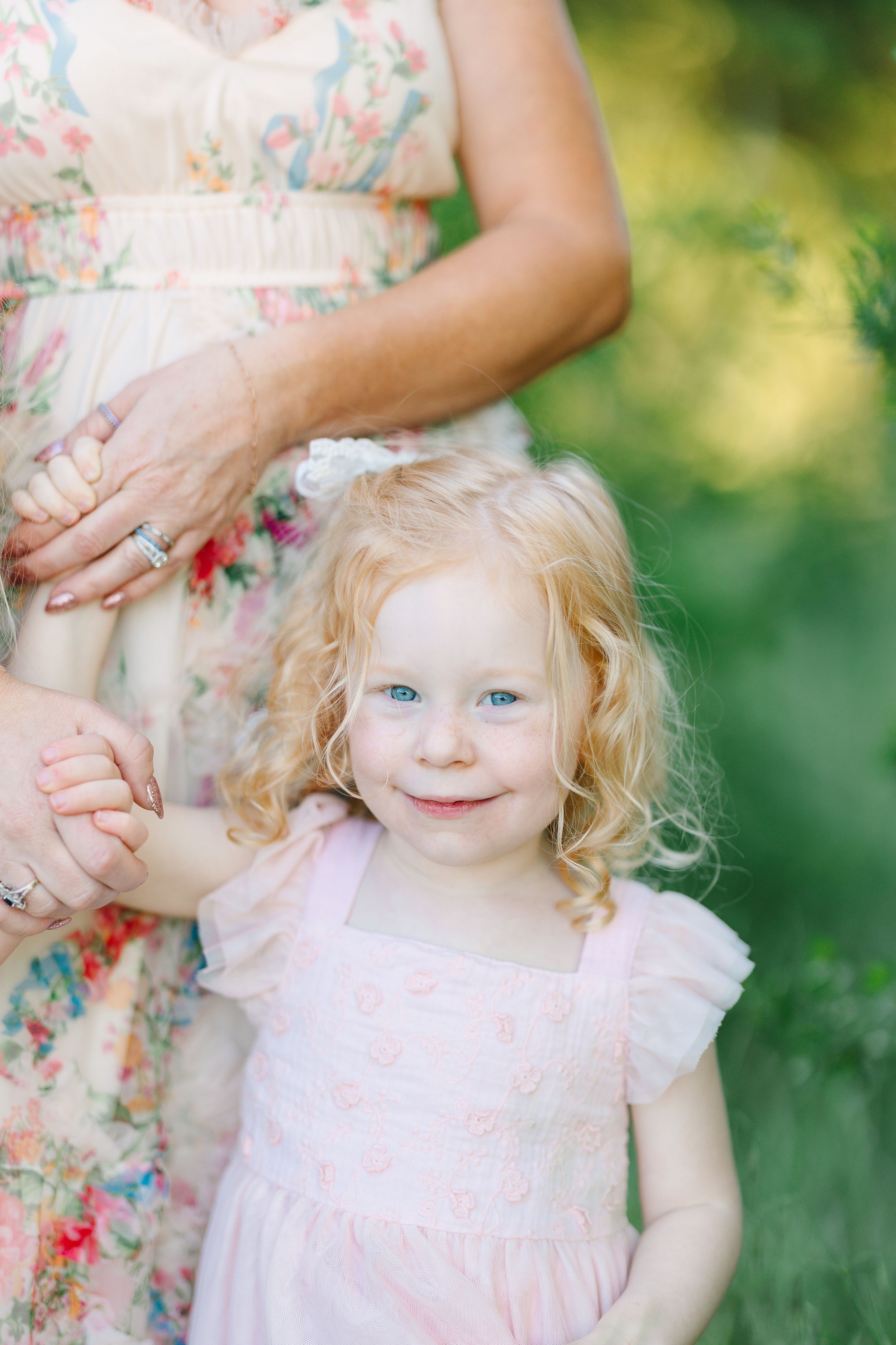 A curly-haired blonde toddler girl with blue eyes in a pink dress holds hands with a woman in a floral dress outdoors.