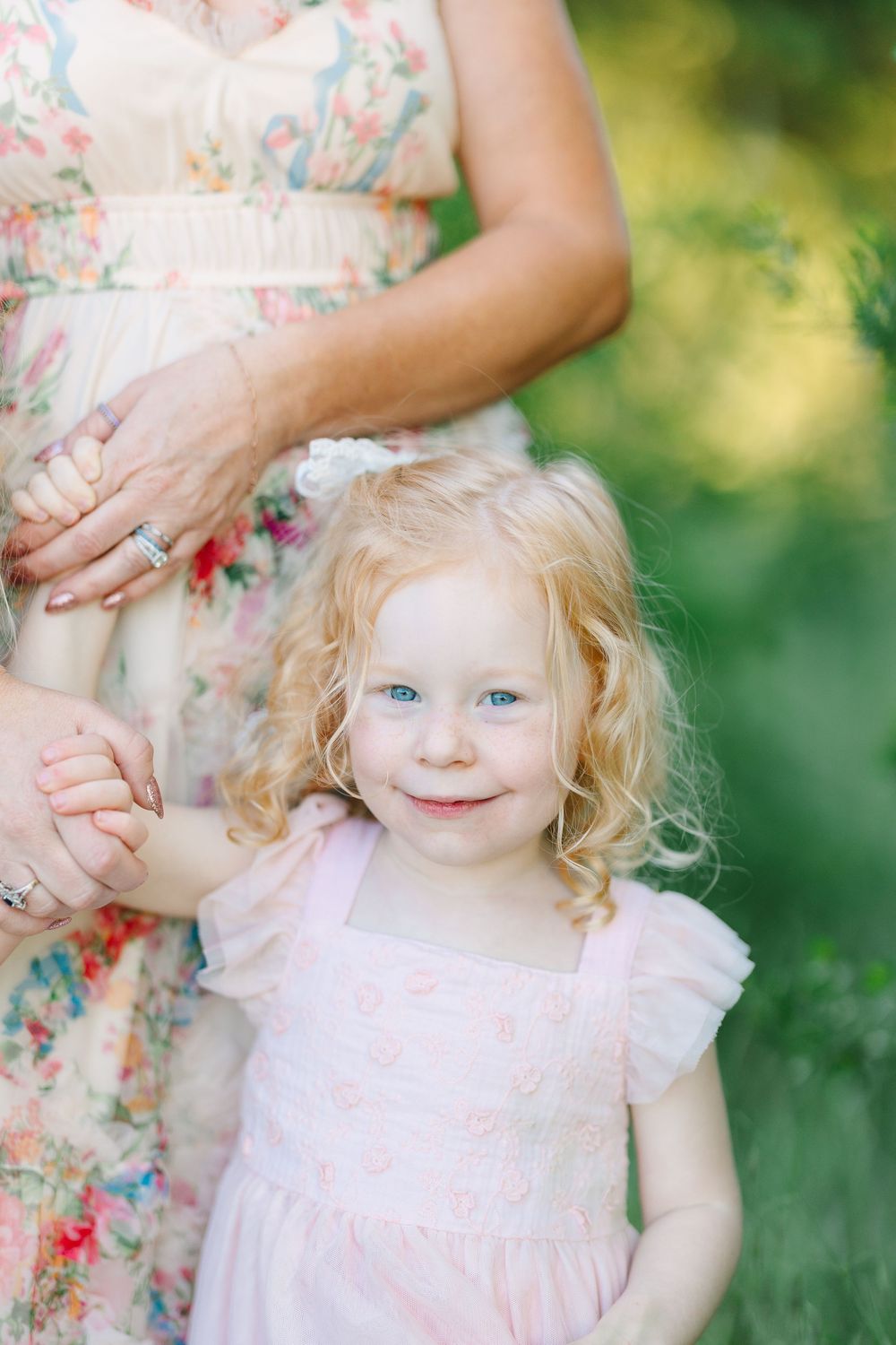 A curly-haired blonde toddler girl with blue eyes in a pink dress holds hands with a woman in a floral dress outdoors.