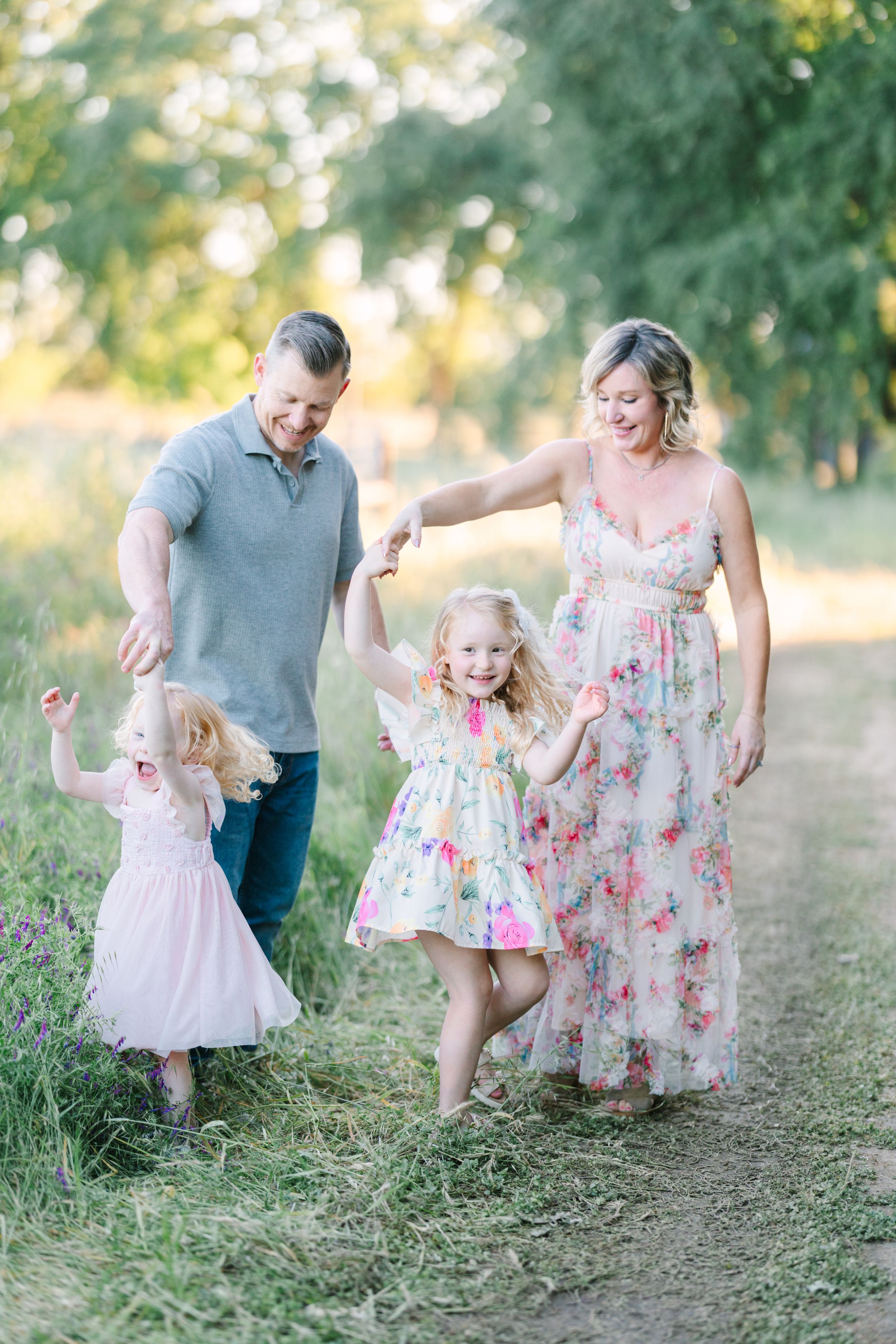 Happy family of four walking outdoors on a sunny path, parents swinging two young girls in floral dresses.
