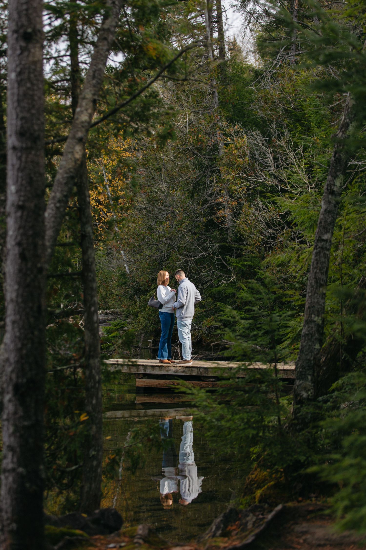 A couple stands together on a wooden dock over a calm pond surrounded by evergreen trees, creating a perfect reflection.